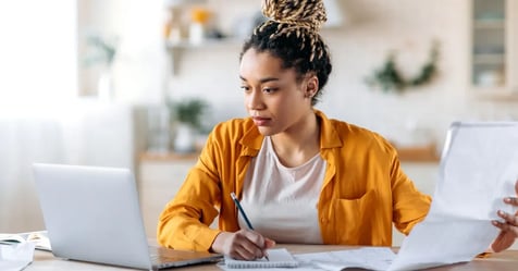 Businesswoman working from home writing on paper, holding a sheet, and looking at her laptop
