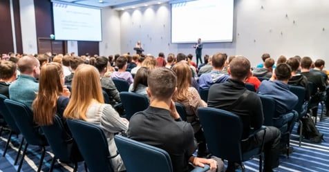 Conference attendees sitting in chairs looking at the stage, listening to the presenter 